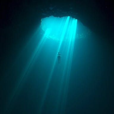 A diver in vast, dark body of water plunging through an opening looking lit by turquoise blue rays through the water