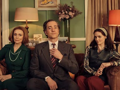 An middle aged woman and man sitting beside a young woman on a sofa, all smartly dressed in a middle class living room