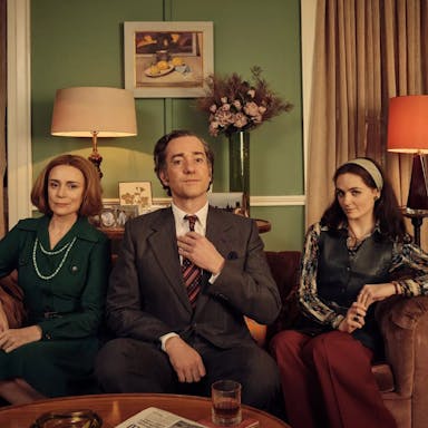 An middle aged woman and man sitting beside a young woman on a sofa, all smartly dressed in a middle class living room