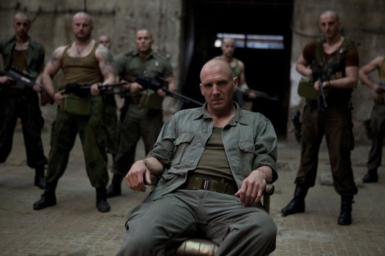A bald white man in military uniform lounges on a chair are lots of bald white men stand behind him holding guns