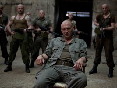 A bald white man in military uniform lounges on a chair are lots of bald white men stand behind him holding guns