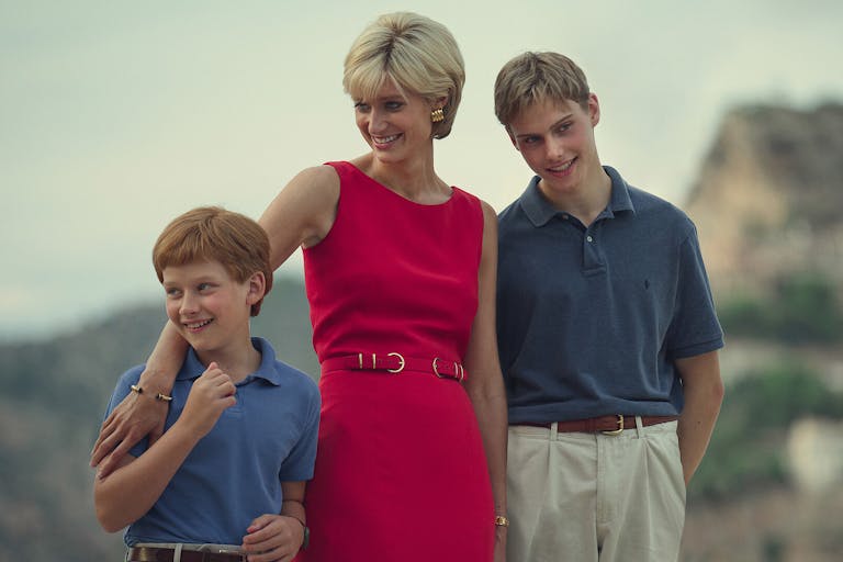 A white woman in a sophisticated hot pink dress with short blonde hair, smiling with her young sons, standing with her arm around the shorter one