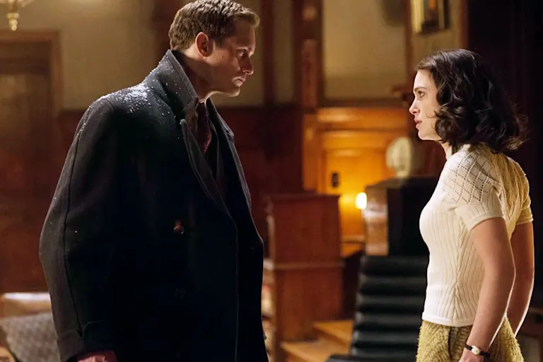 Tall blonde man wearing an overcoat with rain on his shoulders and a young woman in a white top with dark hair look at eachother