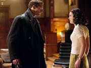 Tall blonde man wearing an overcoat with rain on his shoulders and a young woman in a white top with dark hair look at eachother