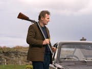 A middle aged white man with brown woollen jacket standing beside a classic car with a shotgun over his shoulder