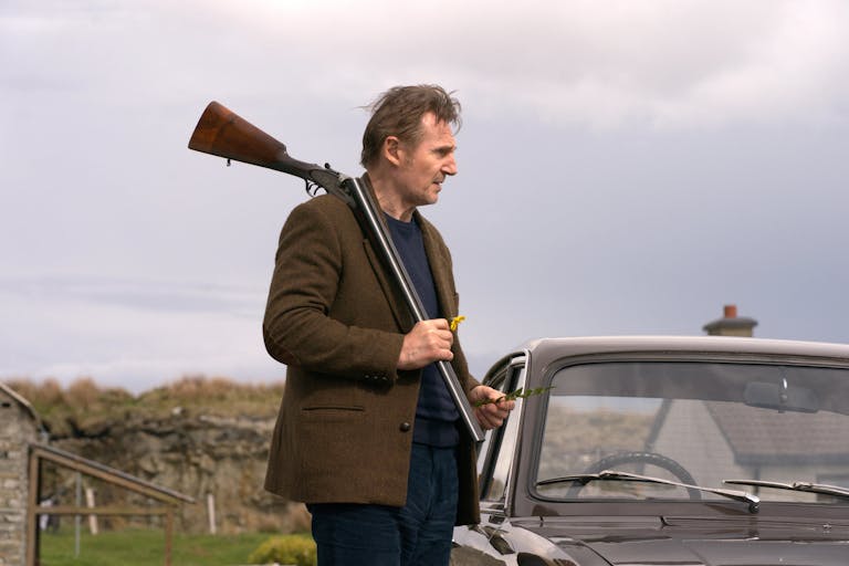 A middle aged white man with brown woollen jacket standing beside a classic car with a shotgun over his shoulder