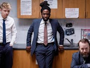 A young Black man in a suit standing at a tired office kitchen, flanked by a standing young white man and a seated older white man
