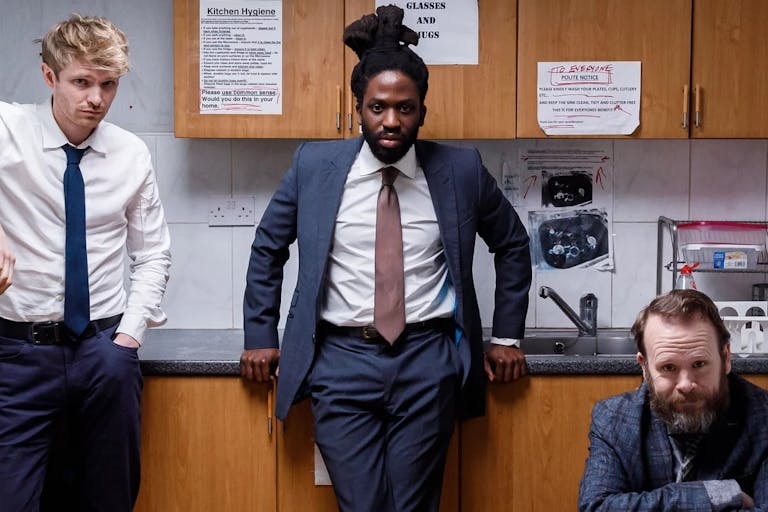 A young Black man in a suit standing at a tired office kitchen, flanked by a standing young white man and a seated older white man