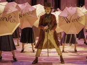 A young white man in along purple coat, brown top hat holding a cane, singing and dancing surrounded by open umbrellas embellished with the word 'Wonka'