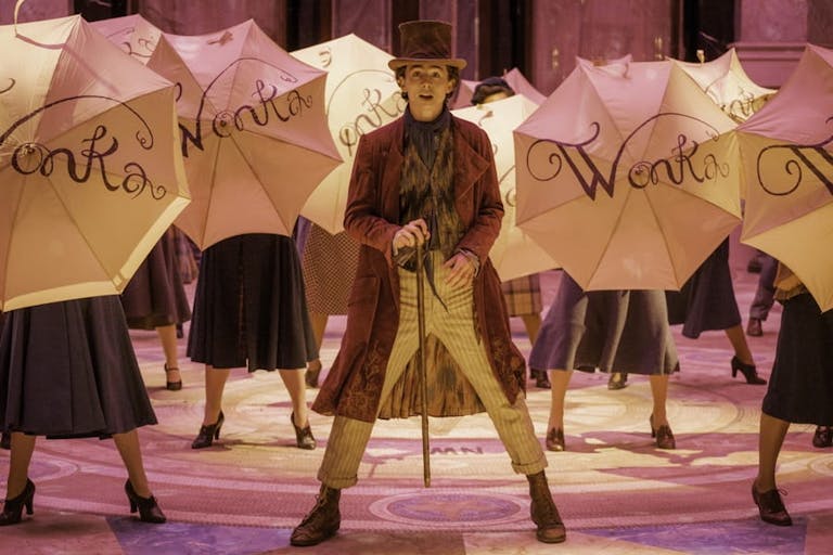 A young white man in along purple coat, brown top hat holding a cane, singing and dancing surrounded by open umbrellas embellished with the word 'Wonka'