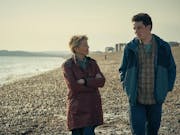 Middle-aged woman with short blonde hair and young man with dark hair walk along a pebbled beach.