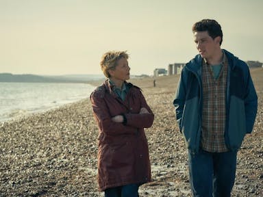 Middle-aged woman with short blonde hair and young man with dark hair walk along a pebbled beach. 