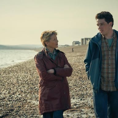 Middle-aged woman with short blonde hair and young man with dark hair walk along a pebbled beach. 