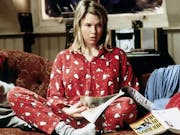 A white blonde woman in red Christmassy penguin pyjamas sits crossed legged on a messy sofa strewn with books and papers
