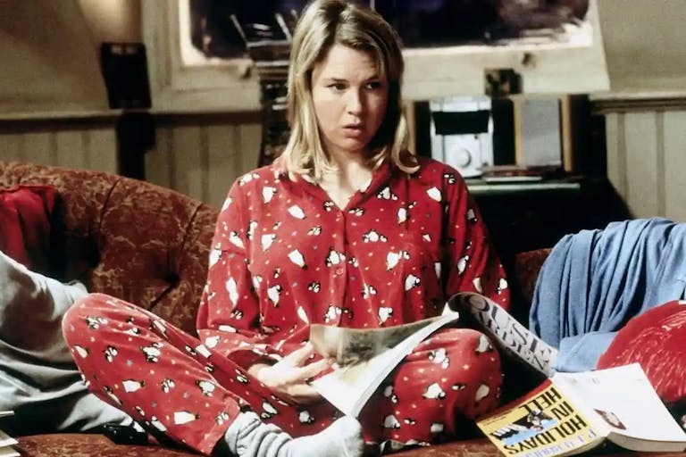 A white blonde woman in red Christmassy penguin pyjamas sits crossed legged on a messy sofa strewn with books and papers