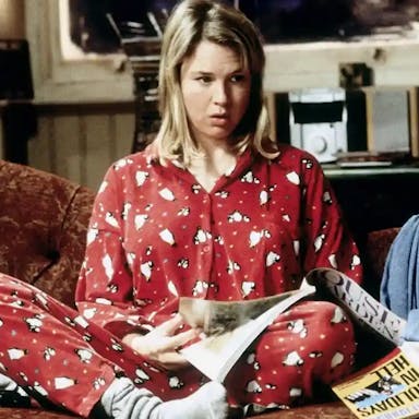 A white blonde woman in red Christmassy penguin pyjamas sits crossed legged on a messy sofa strewn with books and papers