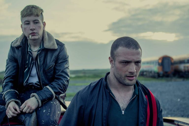 A young white man in leather jacket sits on the roof of a car with a mixed race young Black man walking away with purpose