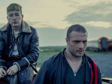 A young white man in leather jacket sits on the roof of a car with a mixed race young Black man walking away with purpose