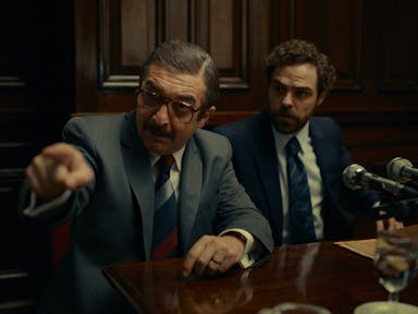 Two men sitting in a courtroom in front of two mics, one pointing their finger 