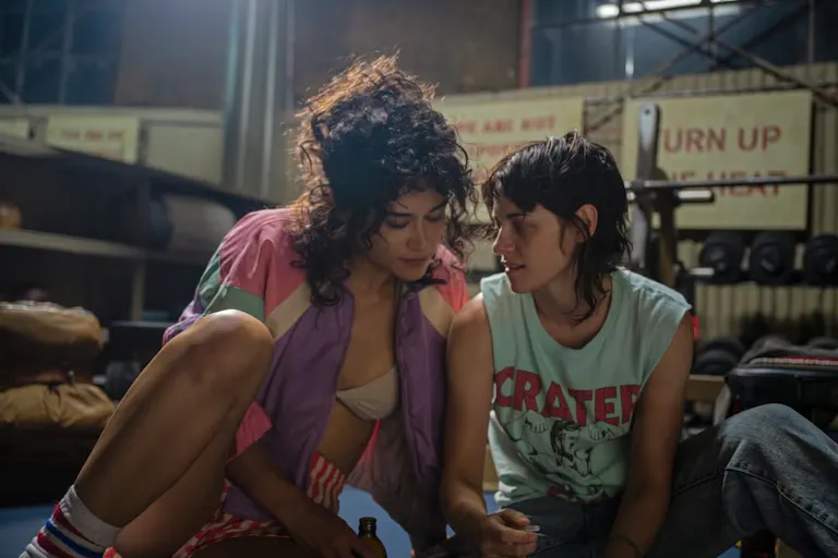 A brown woman wearing a retro 80s jacket, bra and shorts, sitting closely next to a white woman with short dark hair, sleeveless top and baggy jeans in a dated, old school gym