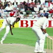 A cricket bowler energetically bowling a red ball at a batter poised to hit, both wearing cricket whites, watched on by spectators in the background