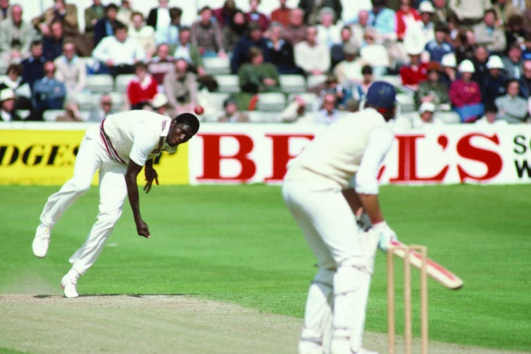 A cricket bowler energetically bowling a red ball at a batter poised to hit, both wearing cricket whites, watched on by spectators in the background