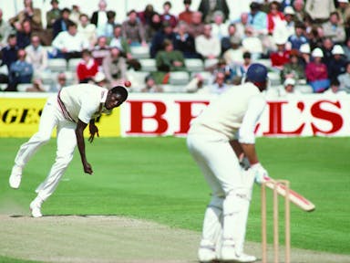 A cricket bowler energetically bowling a red ball at a batter poised to hit, both wearing cricket whites, watched on by spectators in the background