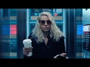 A white woman with long wavy blonde hair and black coat and sunglasses holding a coffee cup looking serious stood in a lift