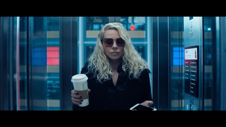 A white woman with long wavy blonde hair and black coat and sunglasses holding a coffee cup looking serious stood in a lift