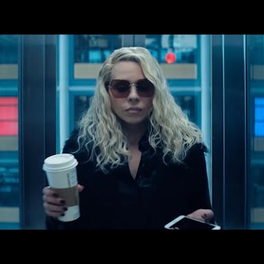 A white woman with long wavy blonde hair and black coat and sunglasses holding a coffee cup looking serious stood in a lift