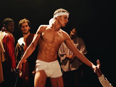 A topless man in a loin cloth and white head band with three men in period clothes behind him and an arm reaching up to him