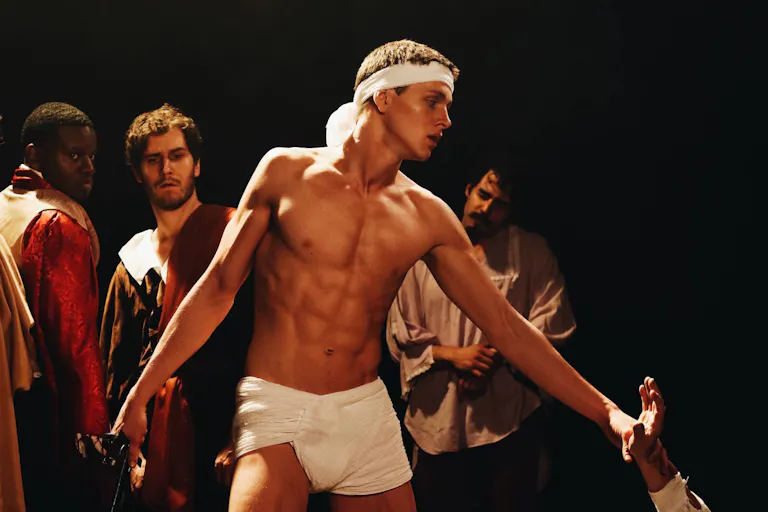A topless man in a loin cloth and white head band with three men in period clothes behind him and an arm reaching up to him