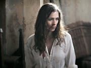 A white woman with long dark hair in a blood stained white shirt and bloody face looking scared
