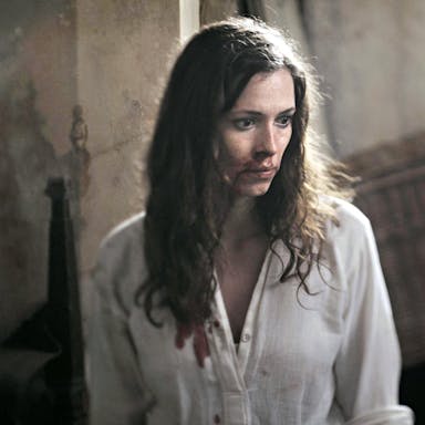 A white woman with long dark hair in a blood stained white shirt and bloody face looking scared
