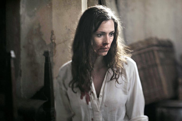 A white woman with long dark hair in a blood stained white shirt and bloody face looking scared