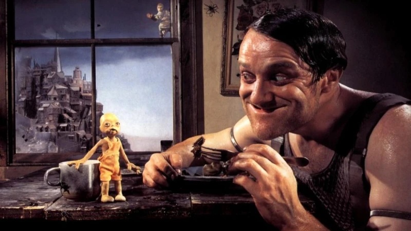A man with a fork in his hand leaning over a table, smiling at a small doll-like creature that is standing on the table. Behind them both is a window showing a dystopian looking area with a tall tower