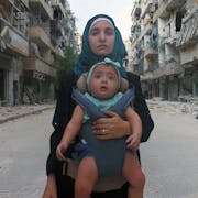 Young woman in a hijab carries her baby in a sling, looking directly at the camera and against a backdrop of a demolished war-torn street