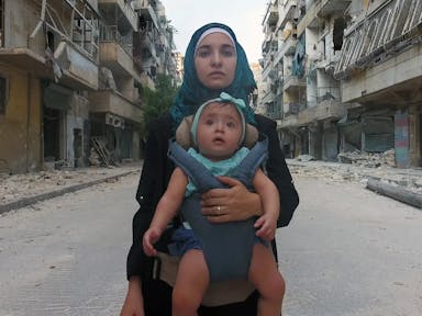 Young woman in a hijab carries her baby in a sling, looking directly at the camera and against a backdrop of a demolished war-torn street