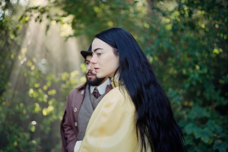 A white woman with long black hair wears a yellow satin dressing gown, stood in a forest with dappled light shining through, a man in a suit stands behind her