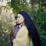 A white woman with long black hair wears a yellow satin dressing gown, stood in a forest with dappled light shining through, a man in a suit stands behind her