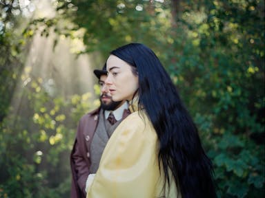 A white woman with long black hair wears a yellow satin dressing gown, stood in a forest with dappled light shining through, a man in a suit stands behind her