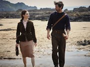 A woman and man in dark clothing walking along an overcast sandy beach
