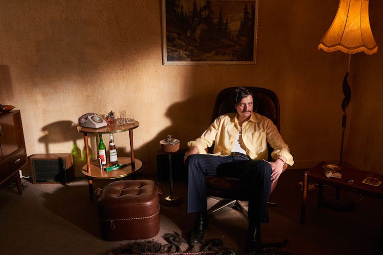 A white man sits in a dim 70's living room in an armchair beside a drinks table with bottles of alcohol and a landline telephone