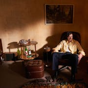 A white man sits in a dim 70's living room in an armchair beside a drinks table with bottles of alcohol and a landline telephone