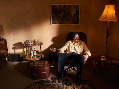 A white man sits in a dim 70's living room in an armchair beside a drinks table with bottles of alcohol and a landline telephone