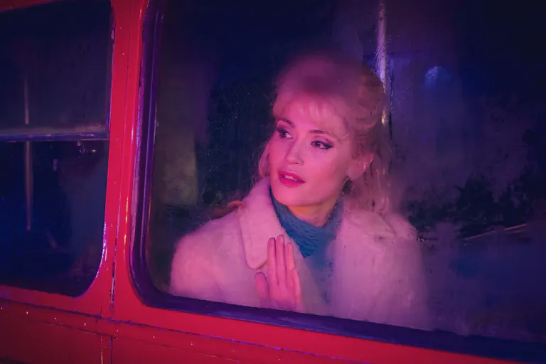 A young white woman with blonde hair in a 60s style looking hopefully out of a red bus's window