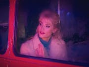 A young white woman with blonde hair in a 60s style looking hopefully out of a red bus's window