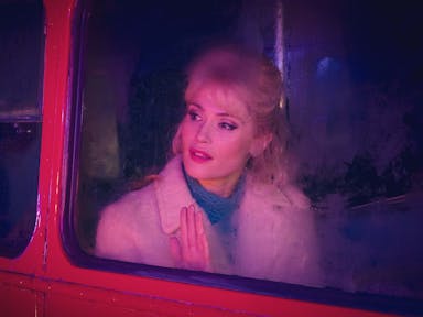 A young white woman with blonde hair in a 60s style looking hopefully out of a red bus's window 