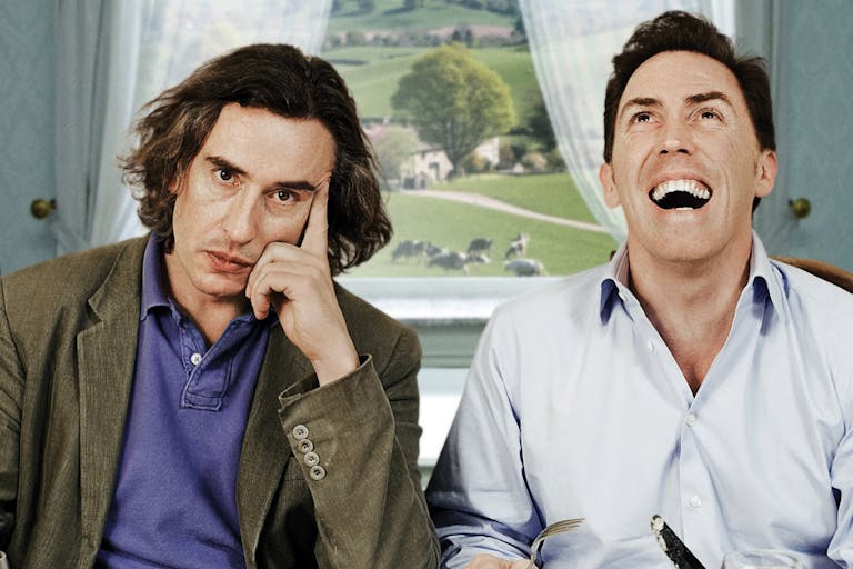 Two middle aged white men sitting at a table with the countryside behind them in the window, one laughing, one looking fed up