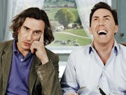 Two middle aged white men sitting at a table with the countryside behind them in the window, one laughing, one looking fed up